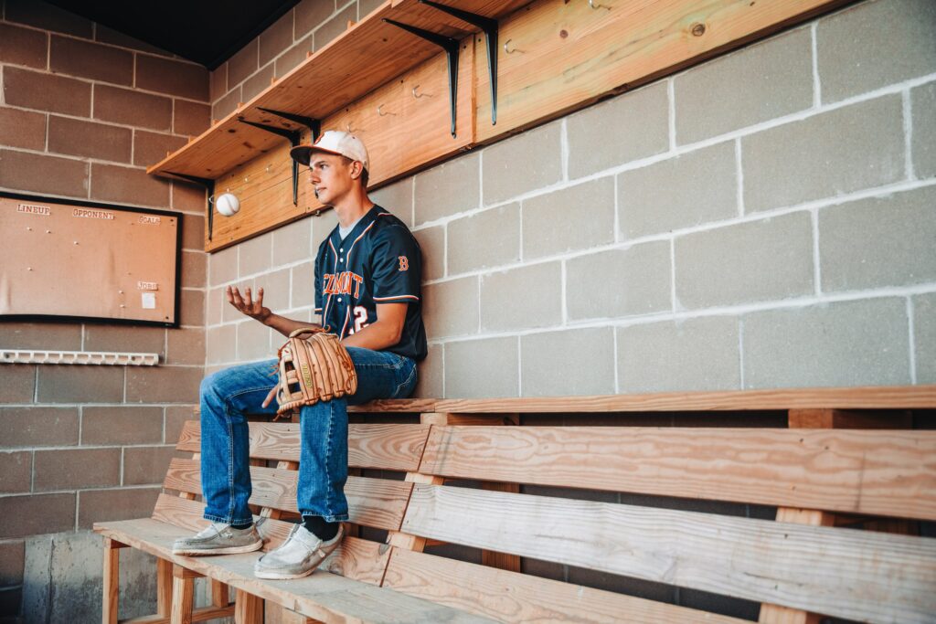 a high school senior guy sitting in the dug out tossing the baseball in the air