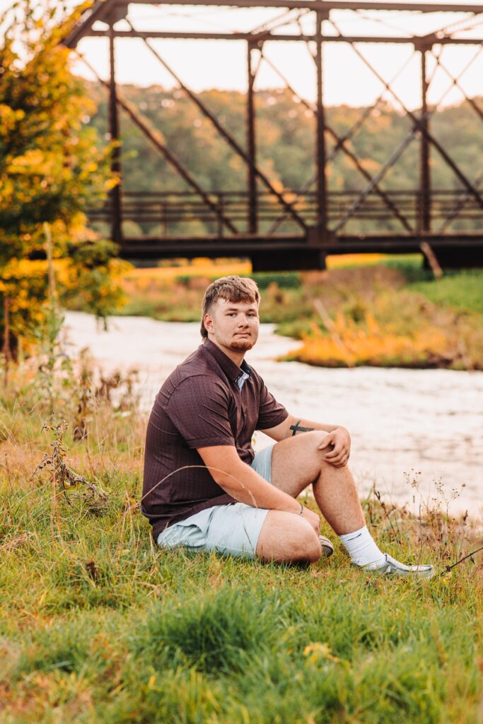 a high school senior guy sitting in the grass with a bridge behind him